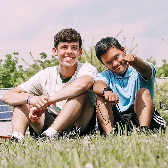 Two young men sitting on grass, smiling and enjoying each other's company. One is wearing a white T-shirt, while the other, in a blue polo, is playfully pointing at the camera. The background features greenery and a bright sky, suggesting an outdoor setting.